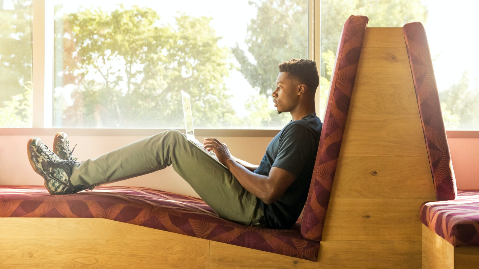 A young male sitting in a modern office space on large padded benches while on his laptop. He is looking at his screen typing, with his feet up on the bench, sitting back comfortably.