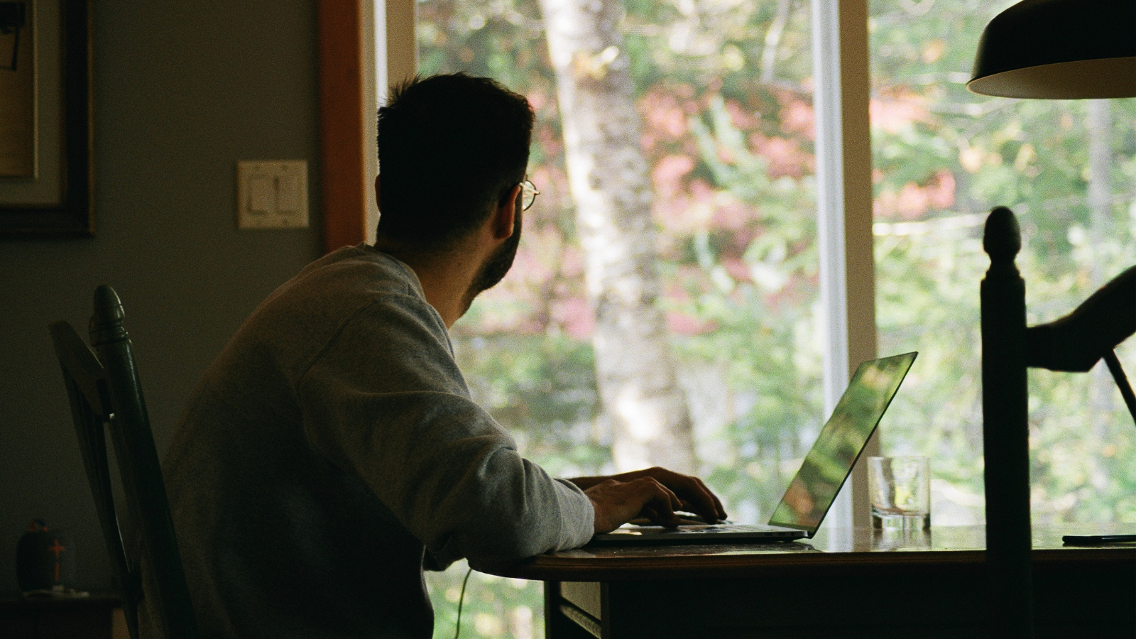 A person sitting at home in their kitchen working on their laptop, but staring away from it, outside their glass door where there is a backyard and trees.